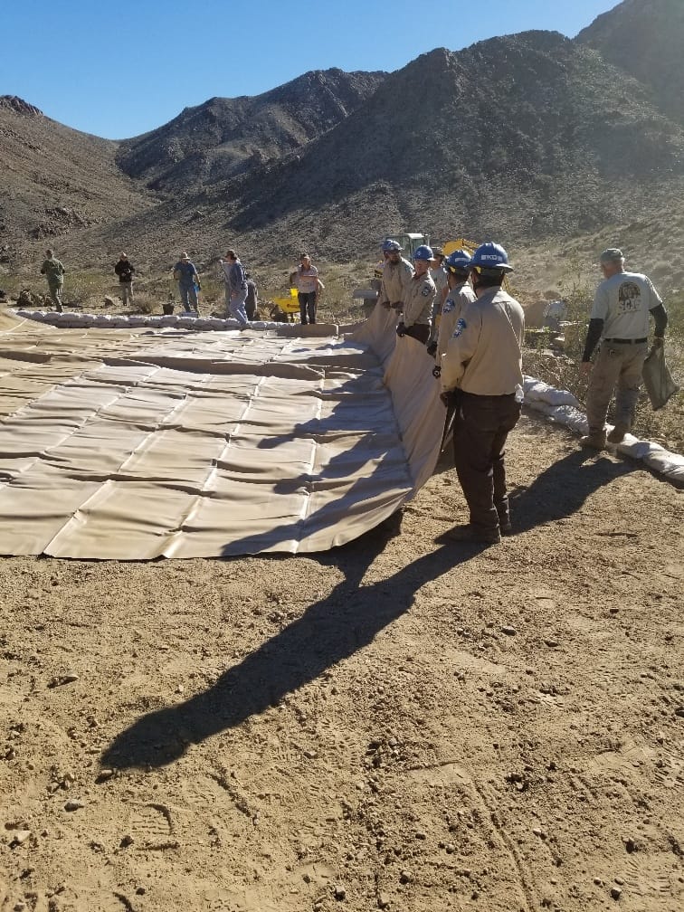 A line of workers in hard hats pulls a large tan liner across the prepared ground