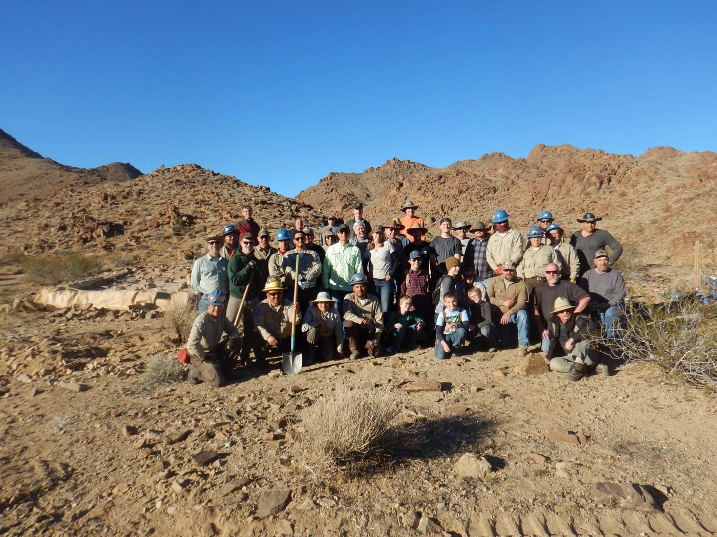 About forty volunteers, several in hard hats, pose together on a rocky desert slope beside the work site