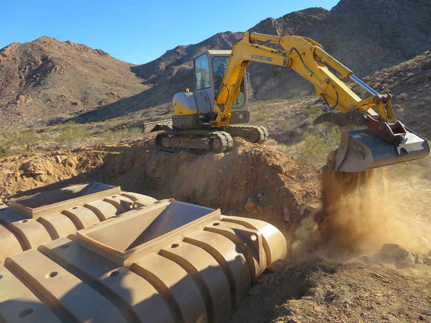 An excavator pushes soil over ribbed plastic water tanks set in a trench