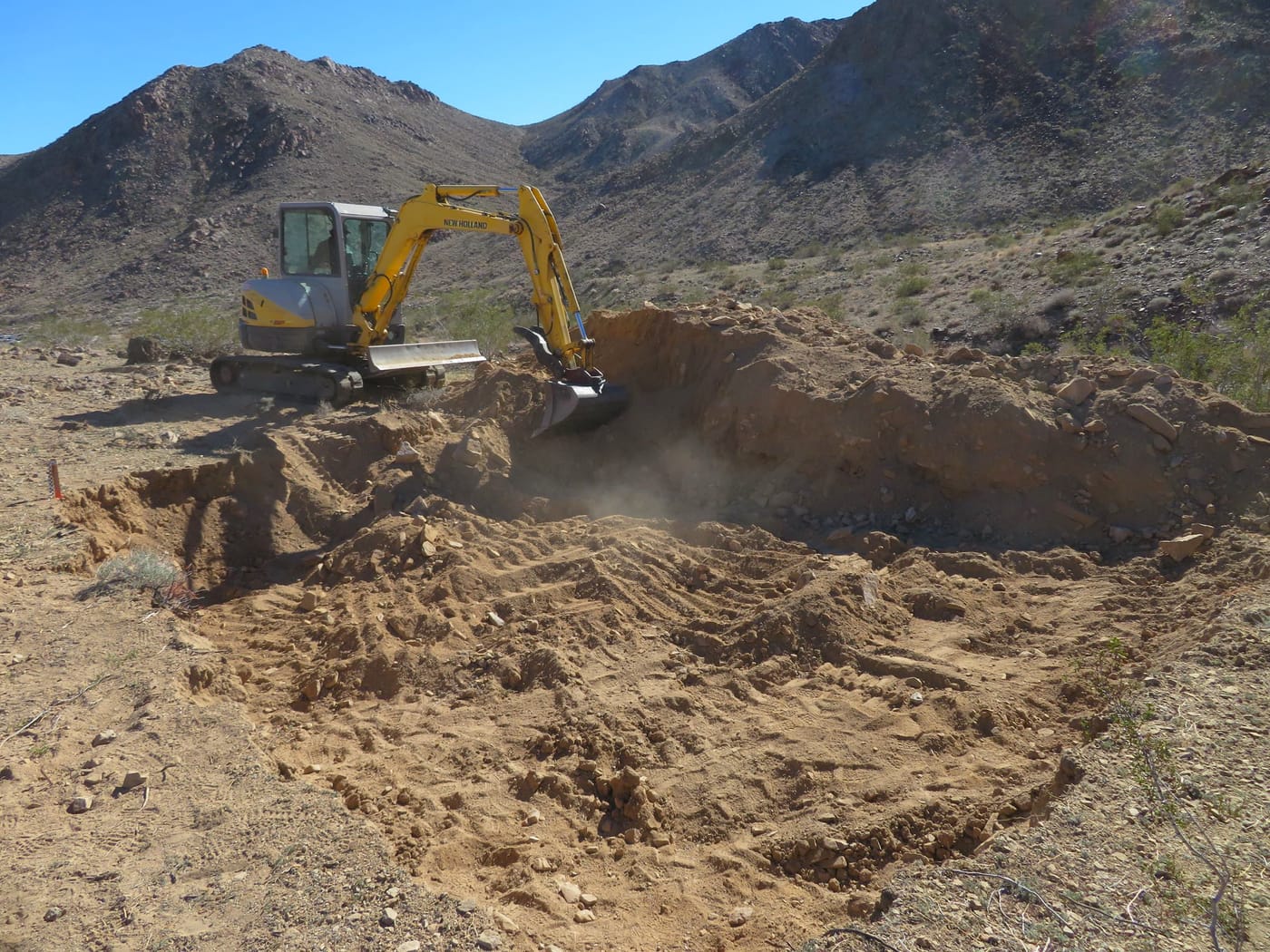 A yellow mini excavator digs a pit in the desert floor below rocky hills
