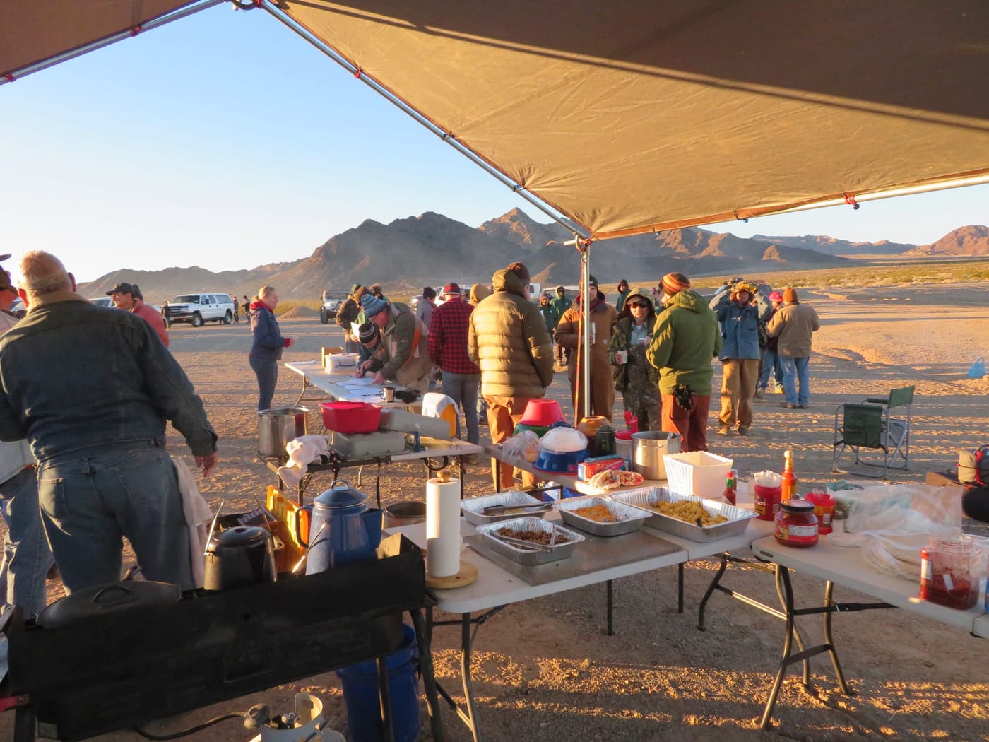 Volunteers in warm jackets gather under a canopy beside tables of food and camp stoves in low desert sun