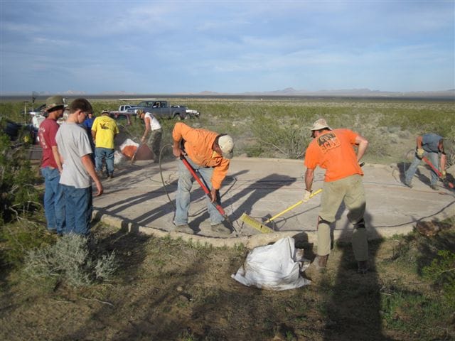 Volunteers sweep and chip at the cracked concrete apron of a desert guzzler as others look on; pickup trucks behind