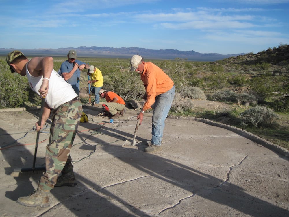 Volunteers with a broom and hand tools work on a guzzler's cracked concrete apron, desert valley and mountains beyond