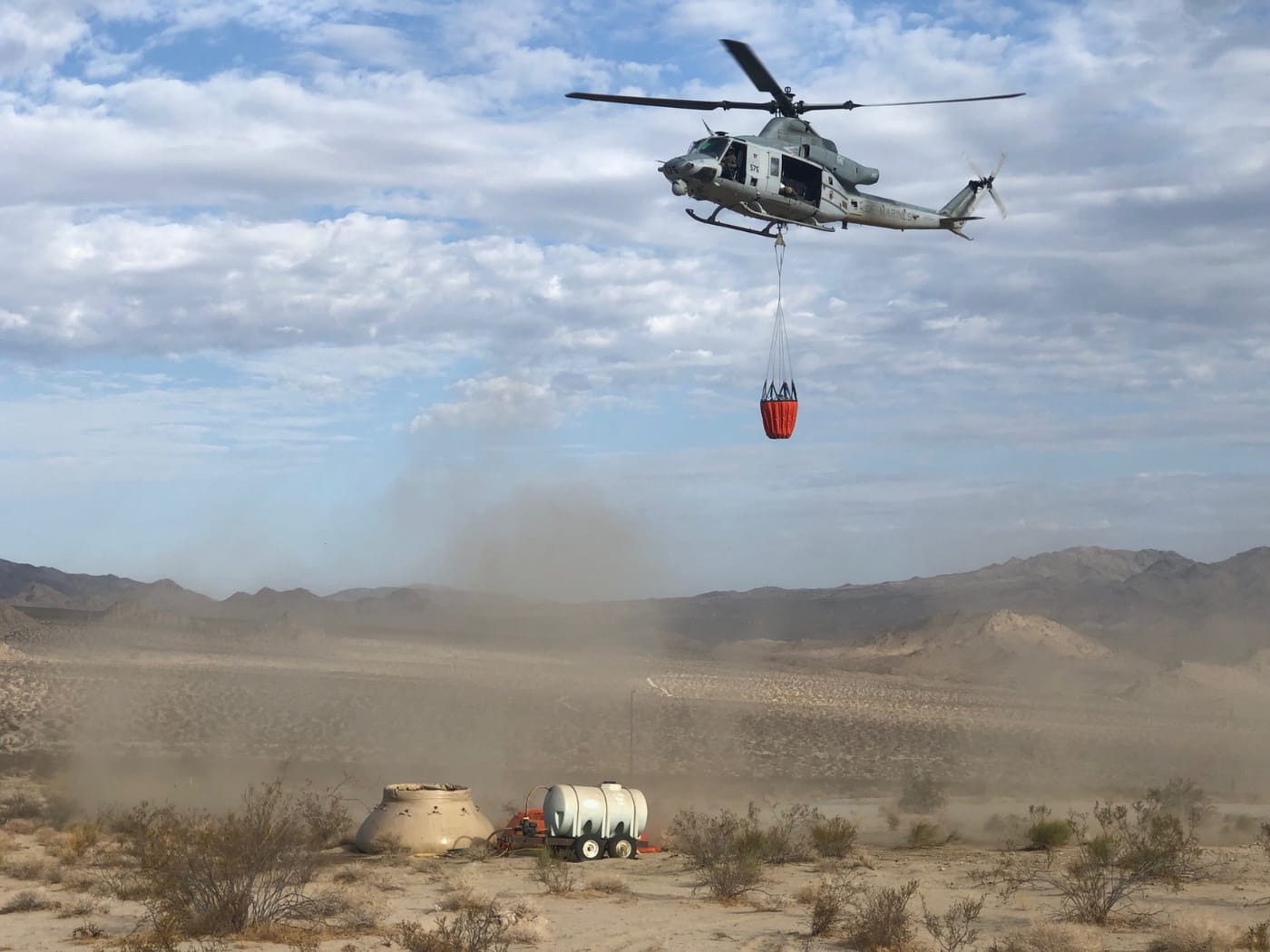 A military helicopter carries an orange water bucket on a long line over open desert; a tank and water trailer sit below