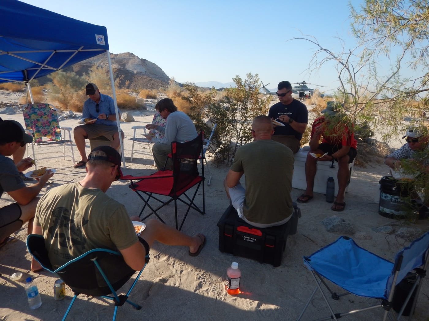 People eat a meal on camp chairs and coolers beside a blue canopy in a sandy wash; a helicopter is parked behind