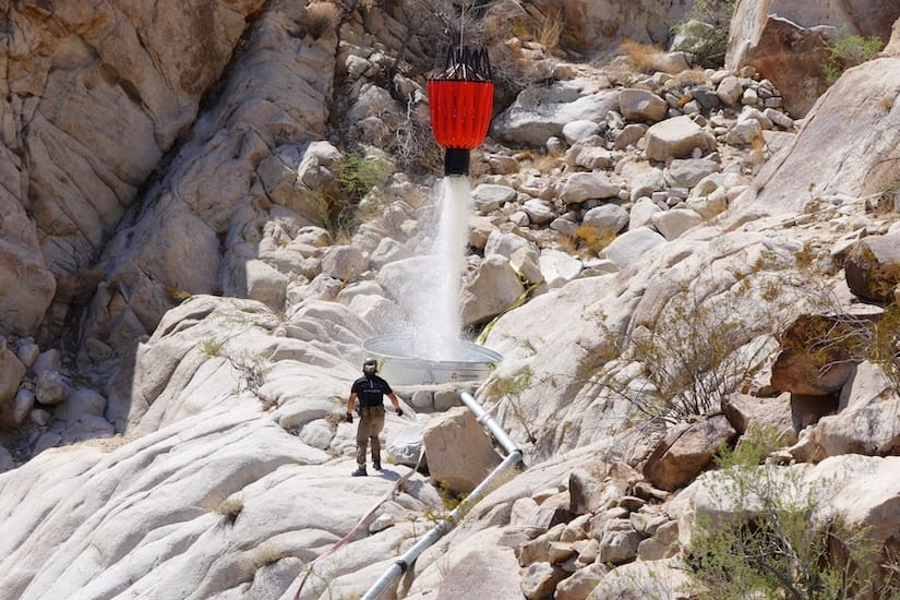 A red helicopter bucket pours water into a round tank set among pale boulders while a crew member watches from the rocks