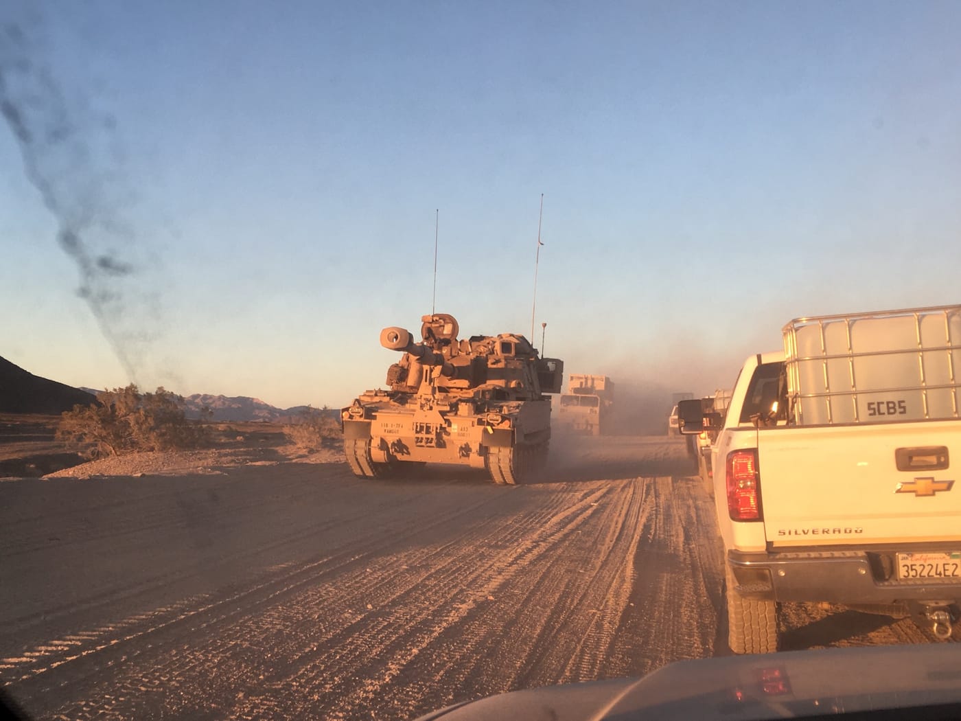 Through a dusty windshield, a tan armored military vehicle passes a white pickup hauling a water tote on a dirt road