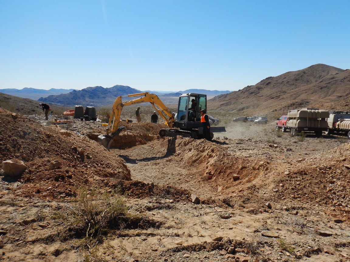 A yellow mini excavator digs a trench on a desert hillside while a trailer loaded with tan water tanks waits nearby