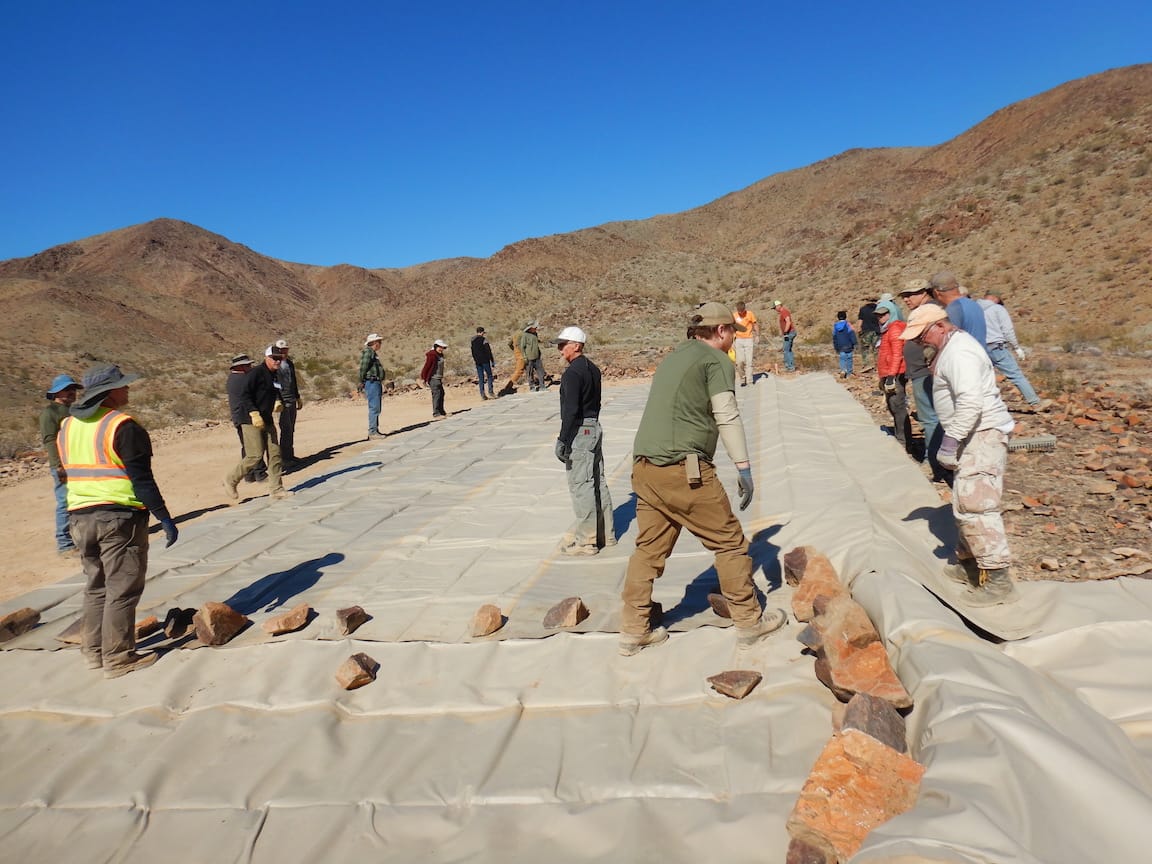 Volunteers walk across a large tan mat laid over cleared desert ground, its edge weighted with rocks