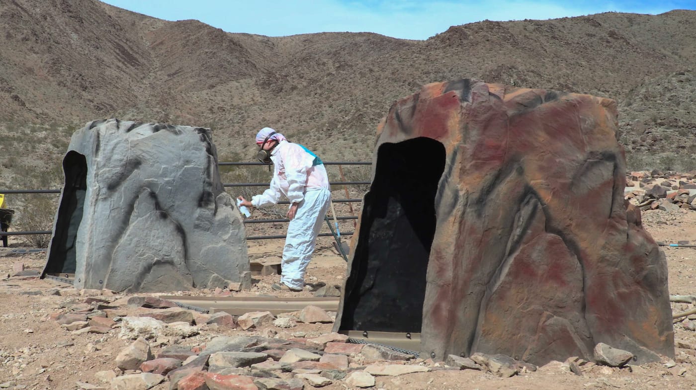 A worker in white coveralls and a respirator spray-paints two rock-shaped drinker housings to match the desert hills