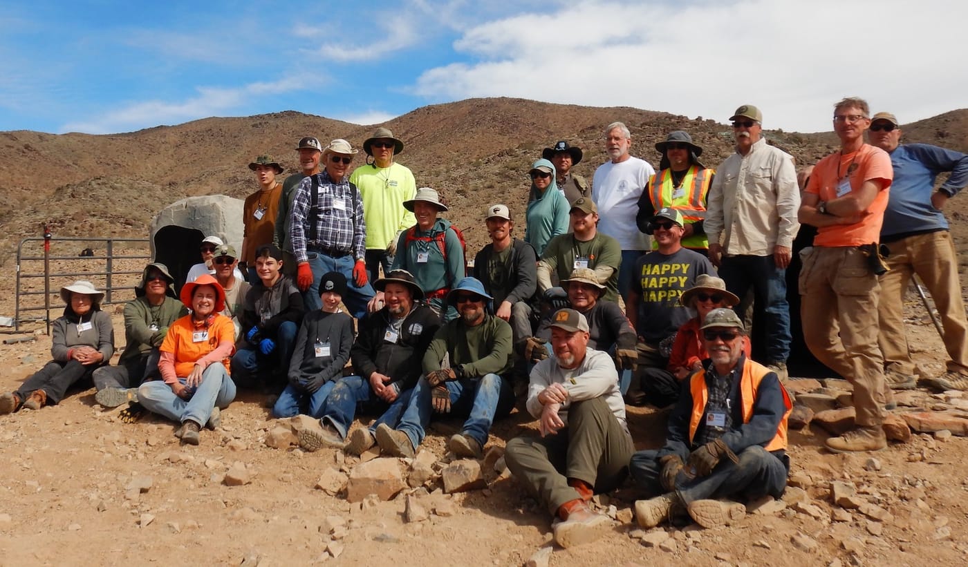 About thirty volunteers pose in rows on a rocky slope, a rock-shaped drinker housing and pipe fence behind them