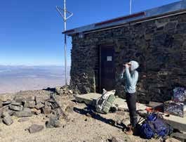 A surveyor glassing with a spotting scope beside the stone hut on White Mountain Peak