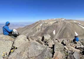 Two people sitting among boulders on a high ridge, looking across to a broad bare mountain streaked with snow
