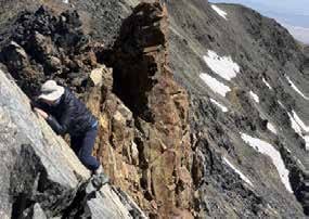 A person in a hat climbing across a narrow, steep rock ridge with snow patches on the slope below