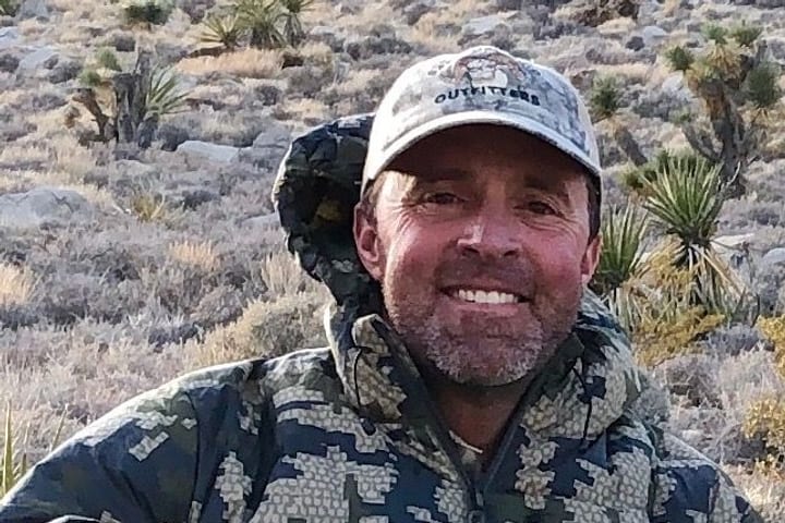 Andrew Pontius smiling in a camouflage jacket and cap on a desert hillside